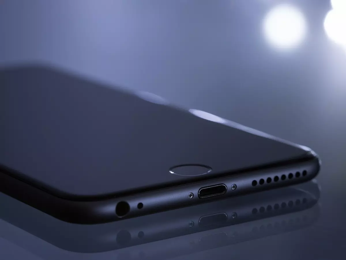 A close-up of a black smartphone lying on a reflective surface with a blurred background. The phone's bottom edge is in focus, showing the charging port, microphone, and speaker grille. The phone's home button is visible in the center of the image.