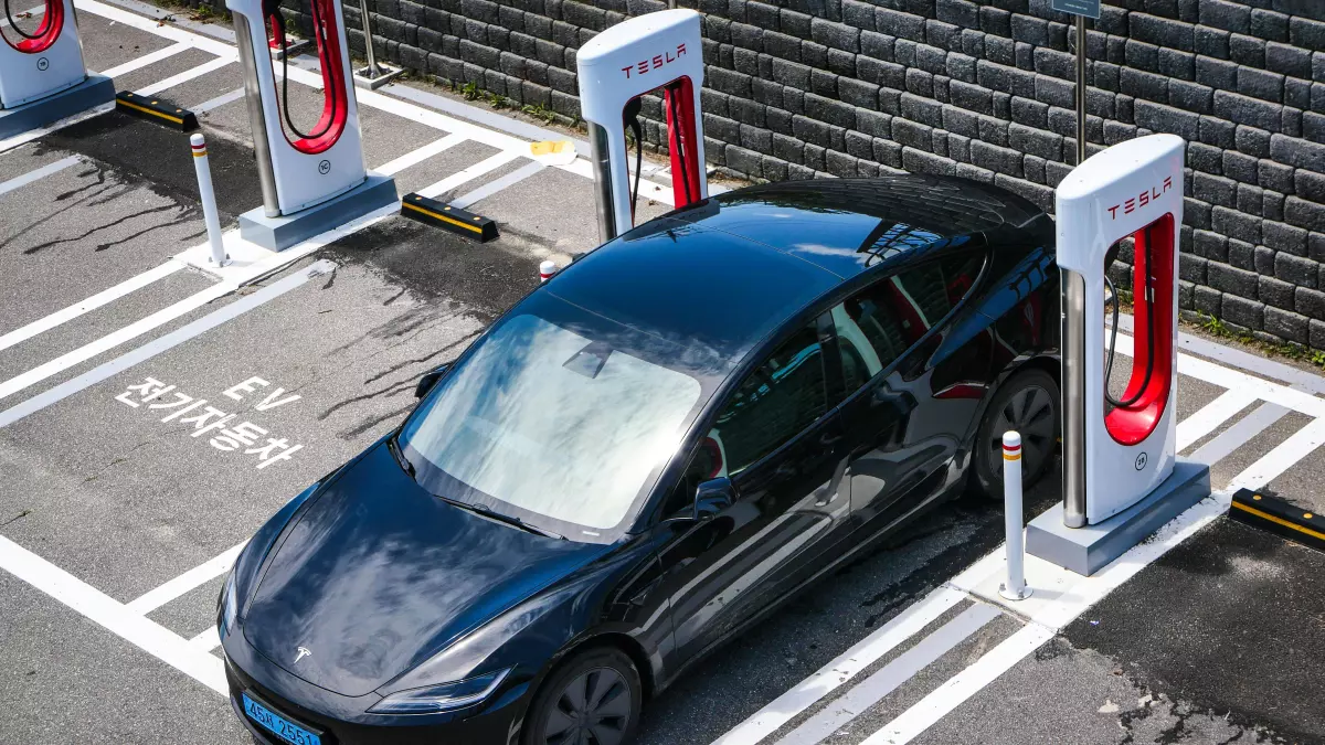 A Tesla Model 3 is charging at a charging station with the charging cable plugged in. The car is parked in a designated charging spot, with a sign indicating that it is reserved for EV charging. The charging station has two charging ports, and the car is connected to one of them.