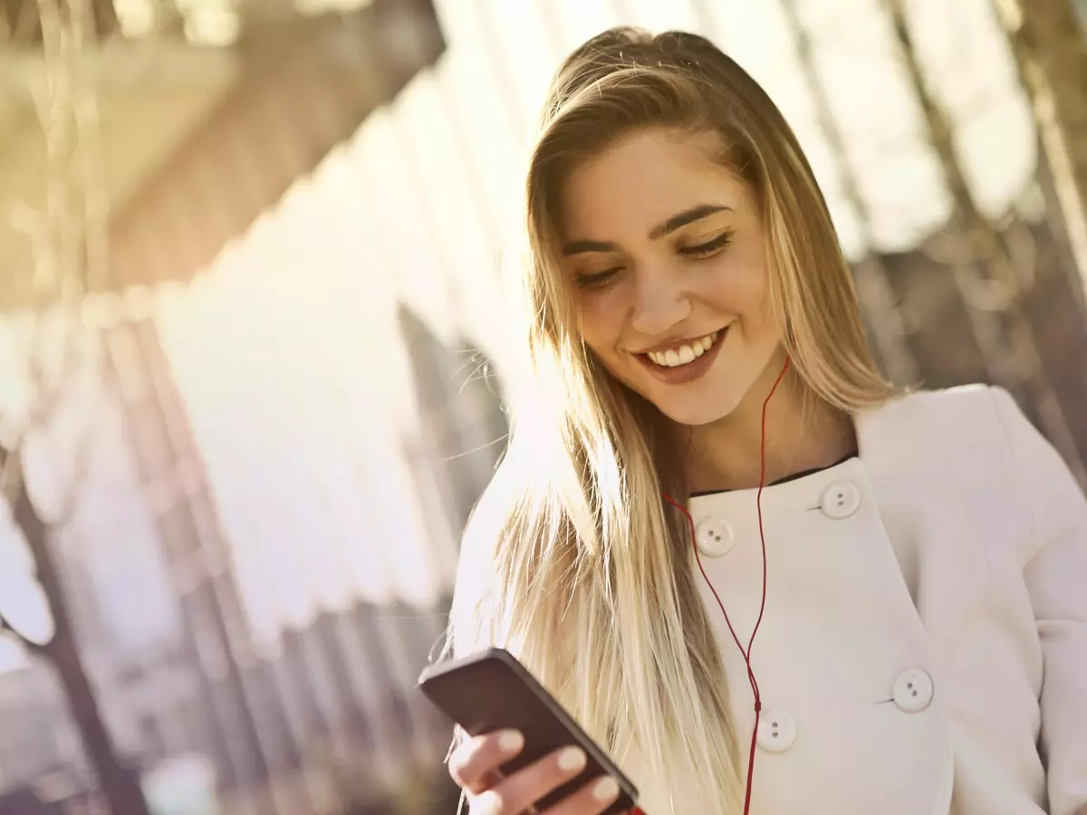 A woman is standing on the street, smiling and looking at her phone.