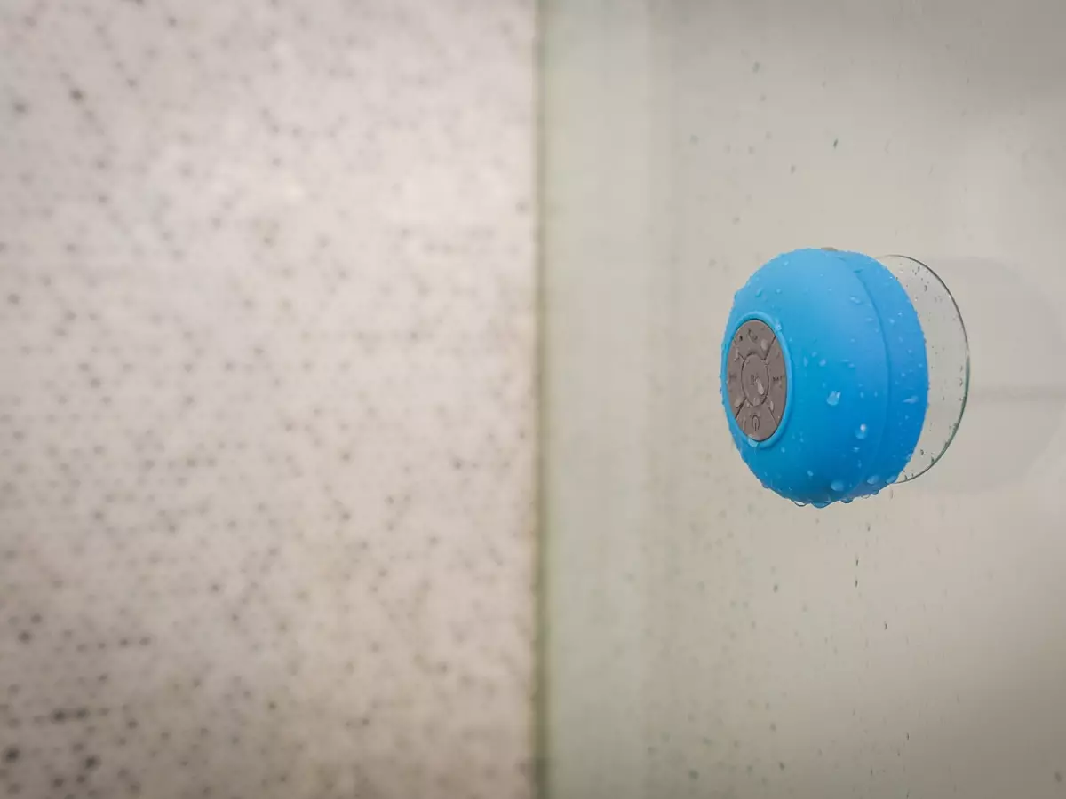 A small blue waterproof speaker with a suction cup is stuck to a shower wall. Water droplets are on the speaker and the wall.