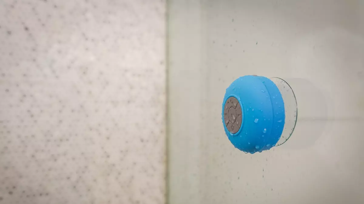 A small blue waterproof speaker with a suction cup is stuck to a shower wall. Water droplets are on the speaker and the wall.