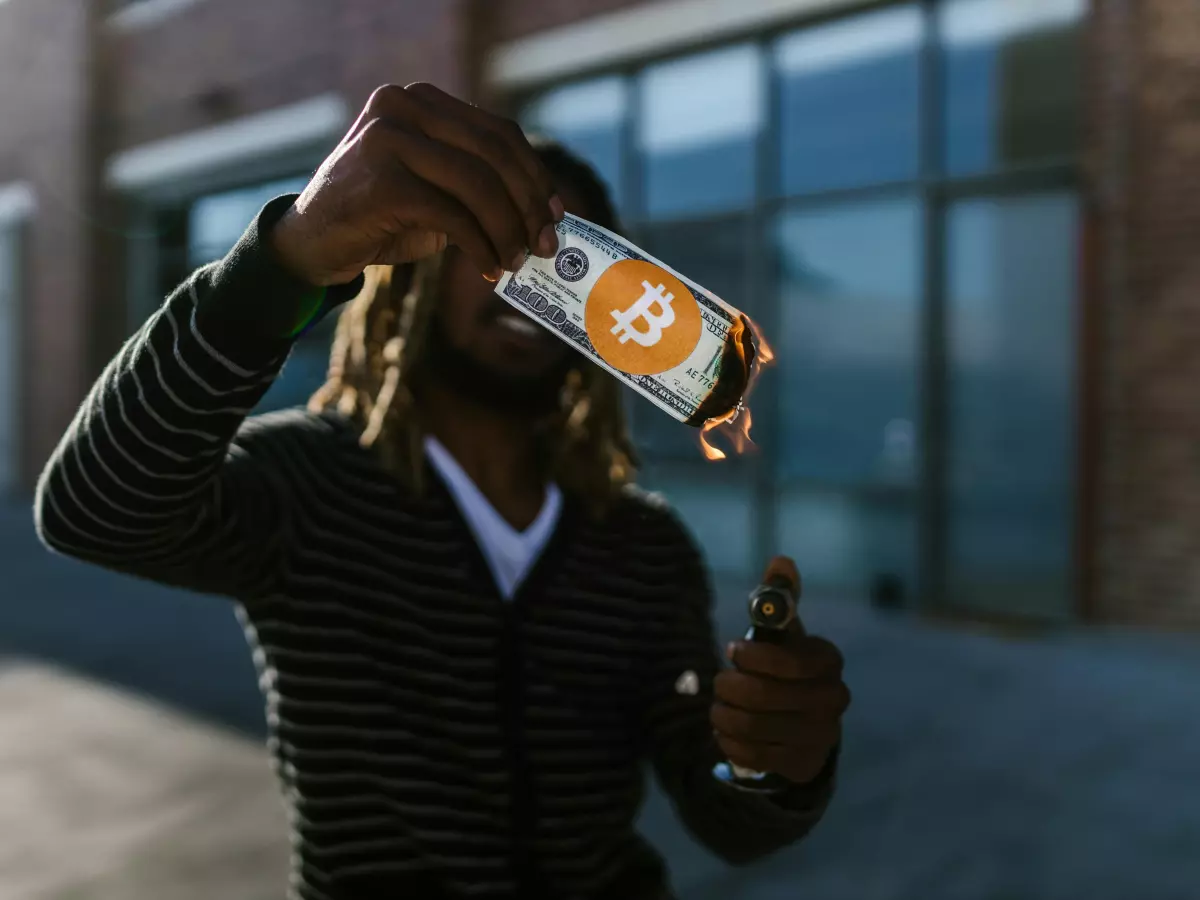 A person in a striped sweater holding a bitcoin coin in front of their face. The bitcoin is a gold color and has the Bitcoin logo on it. The person's face is not visible.