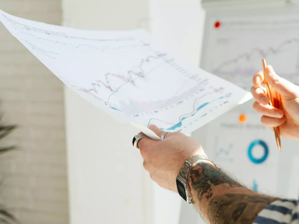 A close-up shot of a person's hands holding and examining a large sheet of paper filled with graphs and charts. The person has tattoos on their arm and is wearing a striped shirt.