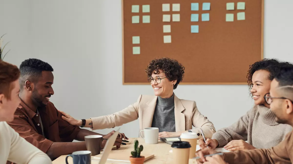 A diverse group of people is sitting around a table, having a friendly meeting, using laptops.