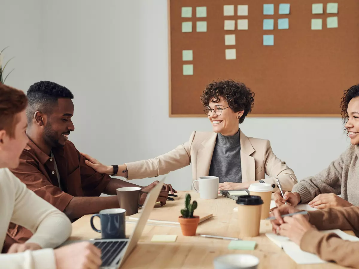 A diverse group of people is sitting around a table, having a friendly meeting, using laptops.