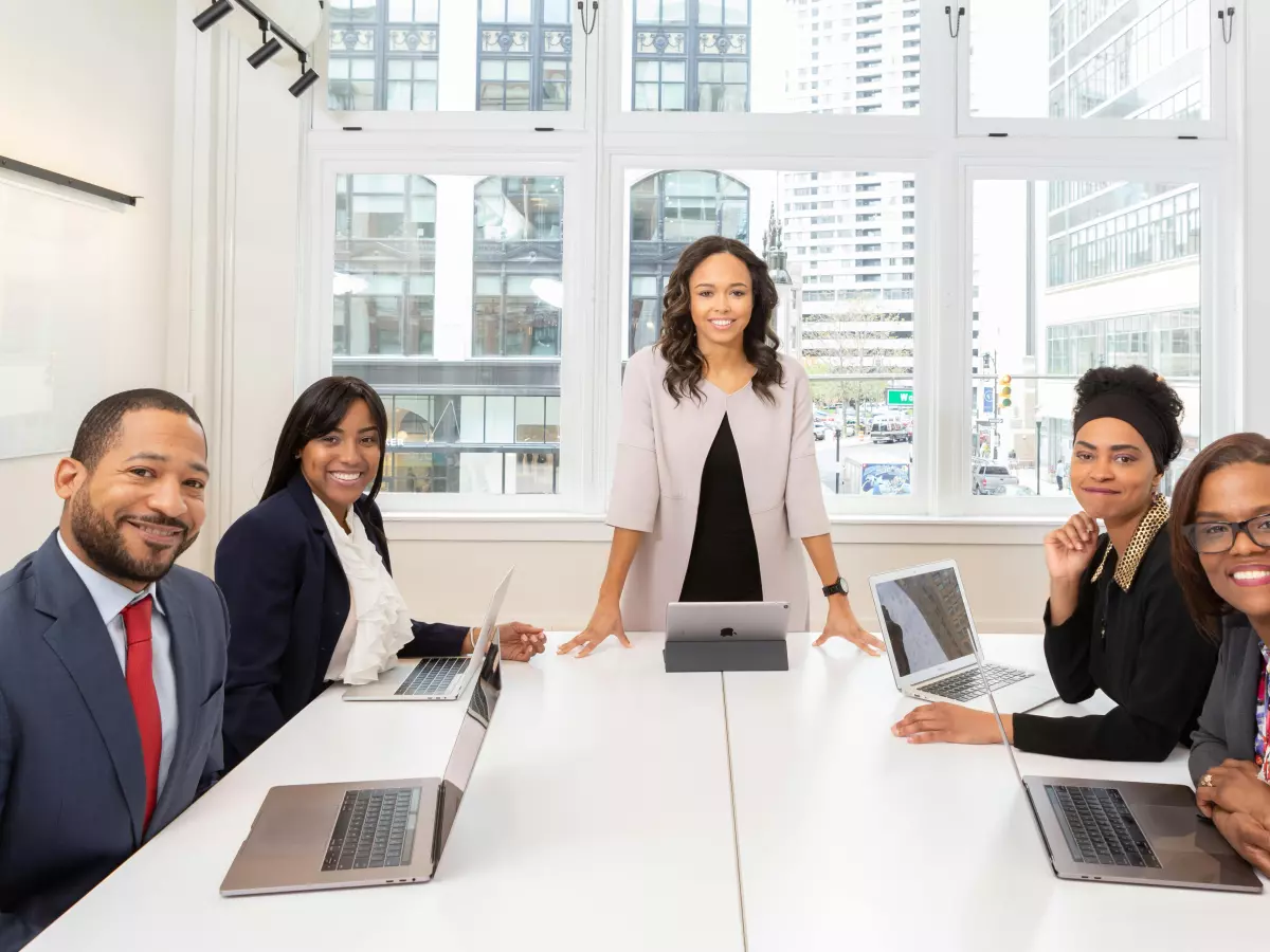 A group of five businesspeople are seated around a table in an office, engaged in a meeting. They are looking at laptops and discussing a topic. The atmosphere is professional and collaborative.