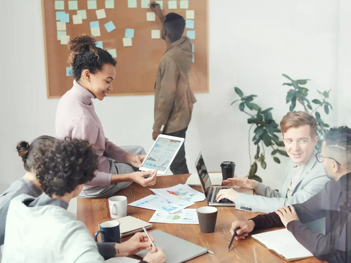 A diverse team of professionals collaborates around a table, using laptops and a whiteboard with sticky notes. 