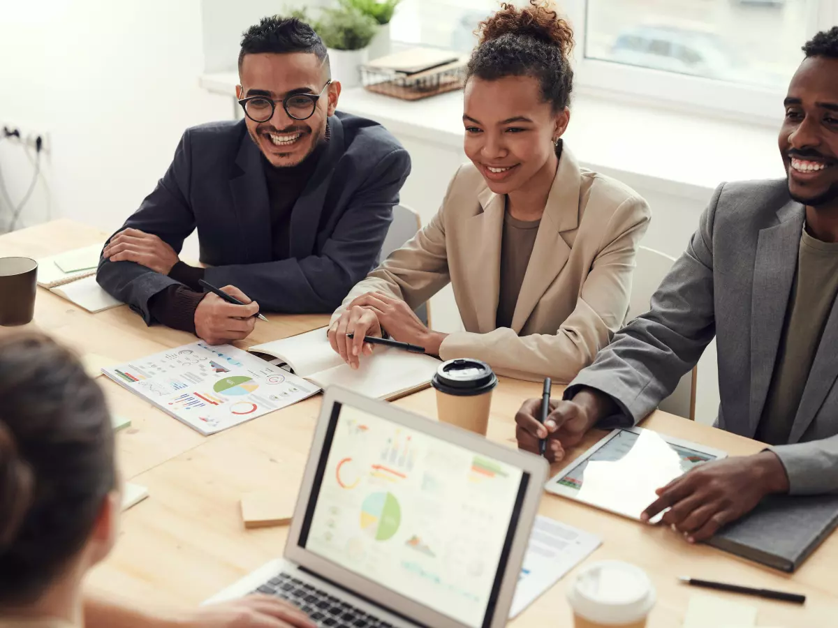 A group of people in business attire are seated around a table in a well-lit office. They are looking at a laptop screen while discussing something. One person is taking notes.