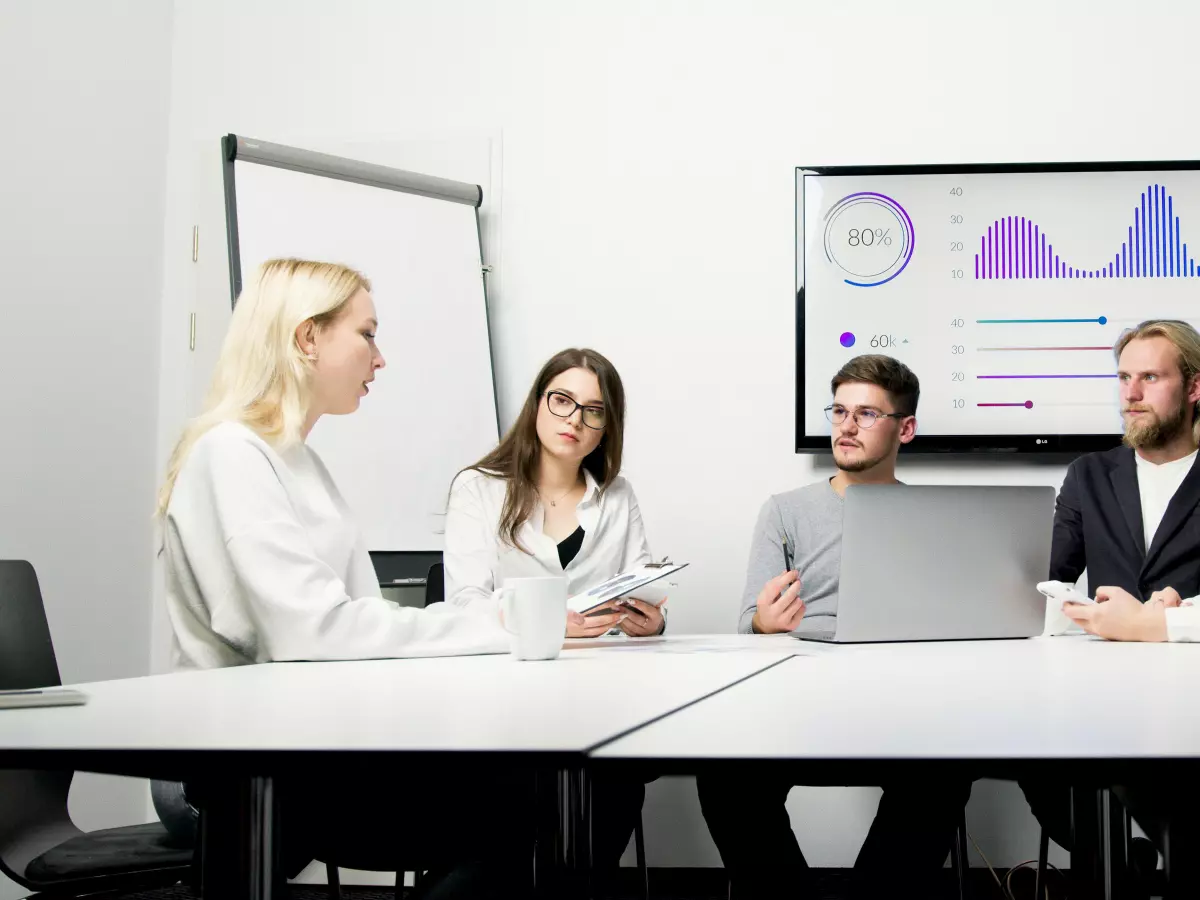 A group of four people sitting around a table in a meeting room. The people are discussing something and there is a laptop computer on the table in front of them. There is a whiteboard behind the people. There is also a large screen with a graph on it behind the people.