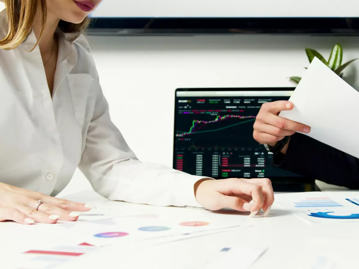 Two people are looking at papers and a screen with graphs. The screen has a graph showing an upward trend. The people are sitting at a table and are talking to each other. The background is a modern office with a window. The dominant color is white.