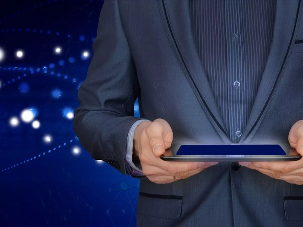 A person in a suit holds a tablet in front of a blue background with abstract network connections and white dots.