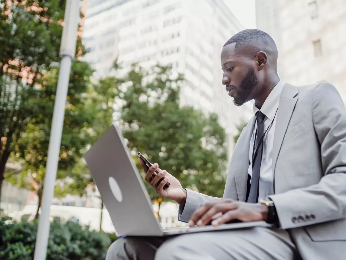 A man in a suit sitting on a bench in a city setting, using a laptop and a smartphone.