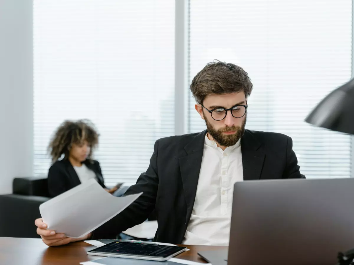 A man in a suit sits at a desk, looking at a laptop, with documents in his hands. A woman in the background is blurred. 