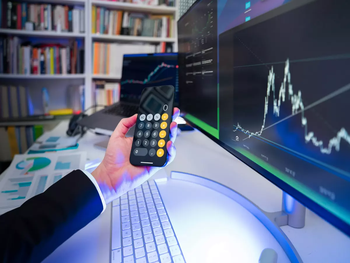 A hand holding a calculator in front of computer screens showing charts