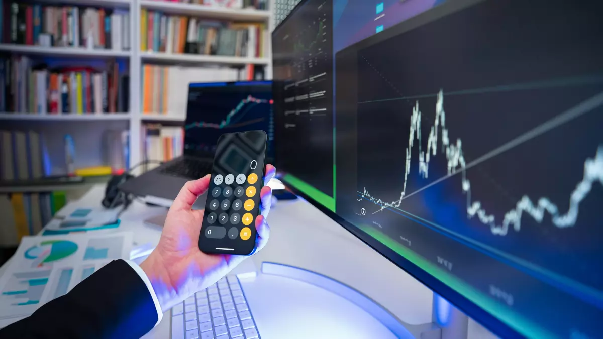 A hand holding a calculator in front of computer screens showing charts