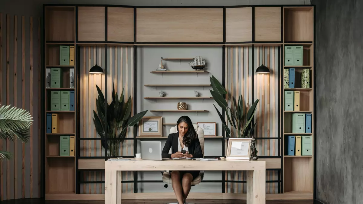 A woman in a suit sits at a large desk in a modern office with built-in bookshelves. The image emphasizes her central role and professional setting.