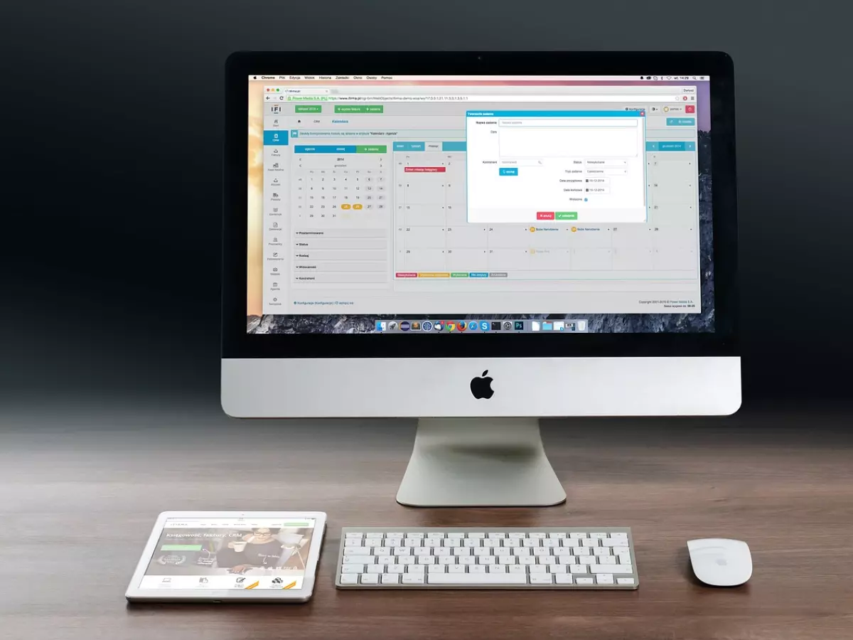 A white Apple iMac desktop computer is on a wooden desk. The screen is showing a calendar app with a notification. There is also a white keyboard and mouse, a white iPad, and a white smartphone on the desk.