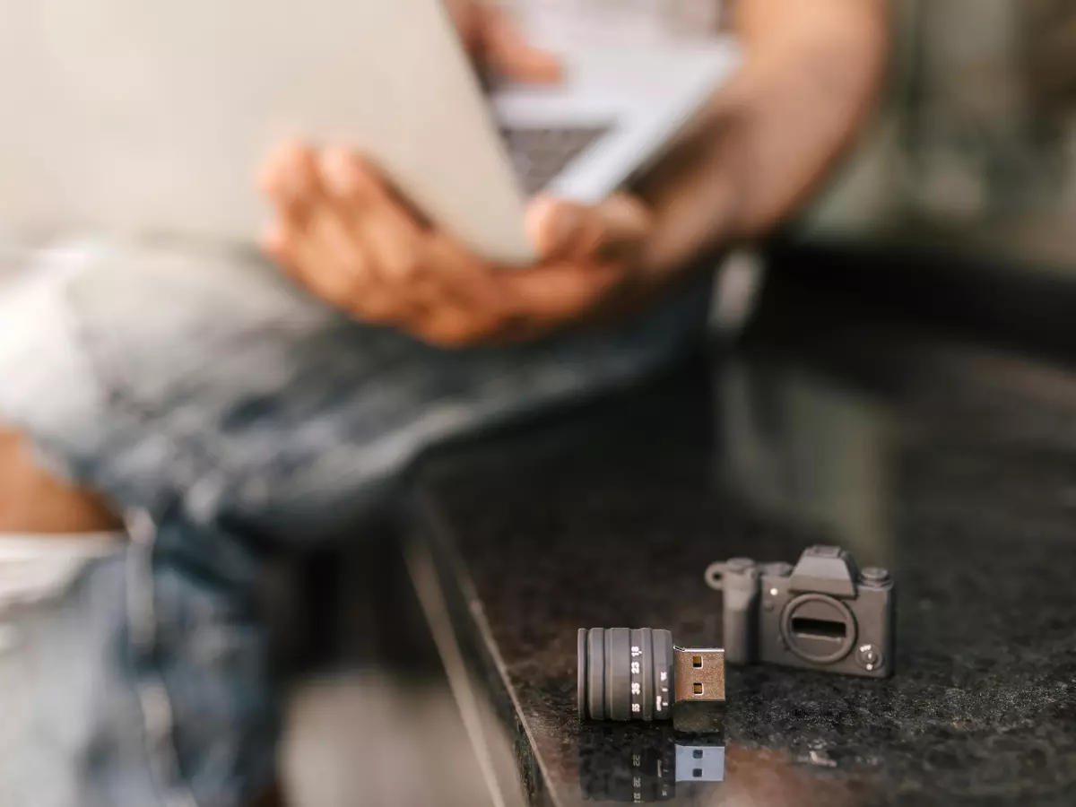 Close-up of a person's hand holding a small device resembling a USB drive. The device is placed on a black surface, and a blurred image of a person sitting on a bench and using a laptop can be seen in the background.