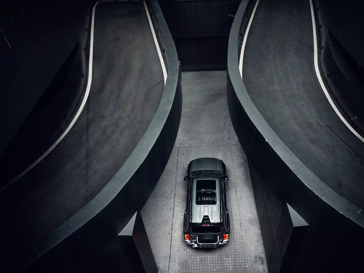 A silver Volvo EX90 SUV is parked in a concrete parking garage. The car is seen from above, and the image is composed in a way that emphasizes the car's sleek design and futuristic features. The concrete garage provides a dramatic backdrop for the car. 