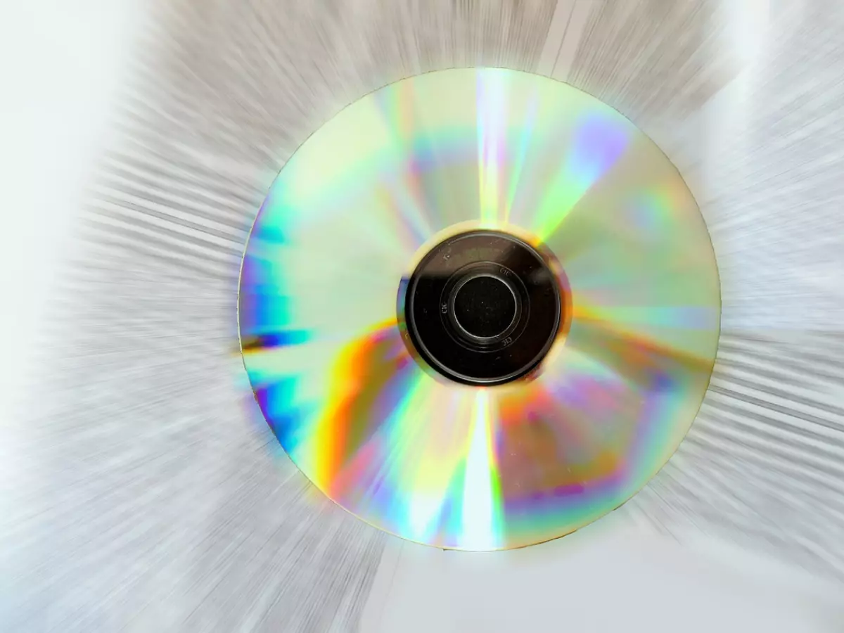 A close-up image of a CD, with light shining through it. The image is taken at a low angle, creating a sense of depth and perspective. The CD is in focus, while the background is blurred.