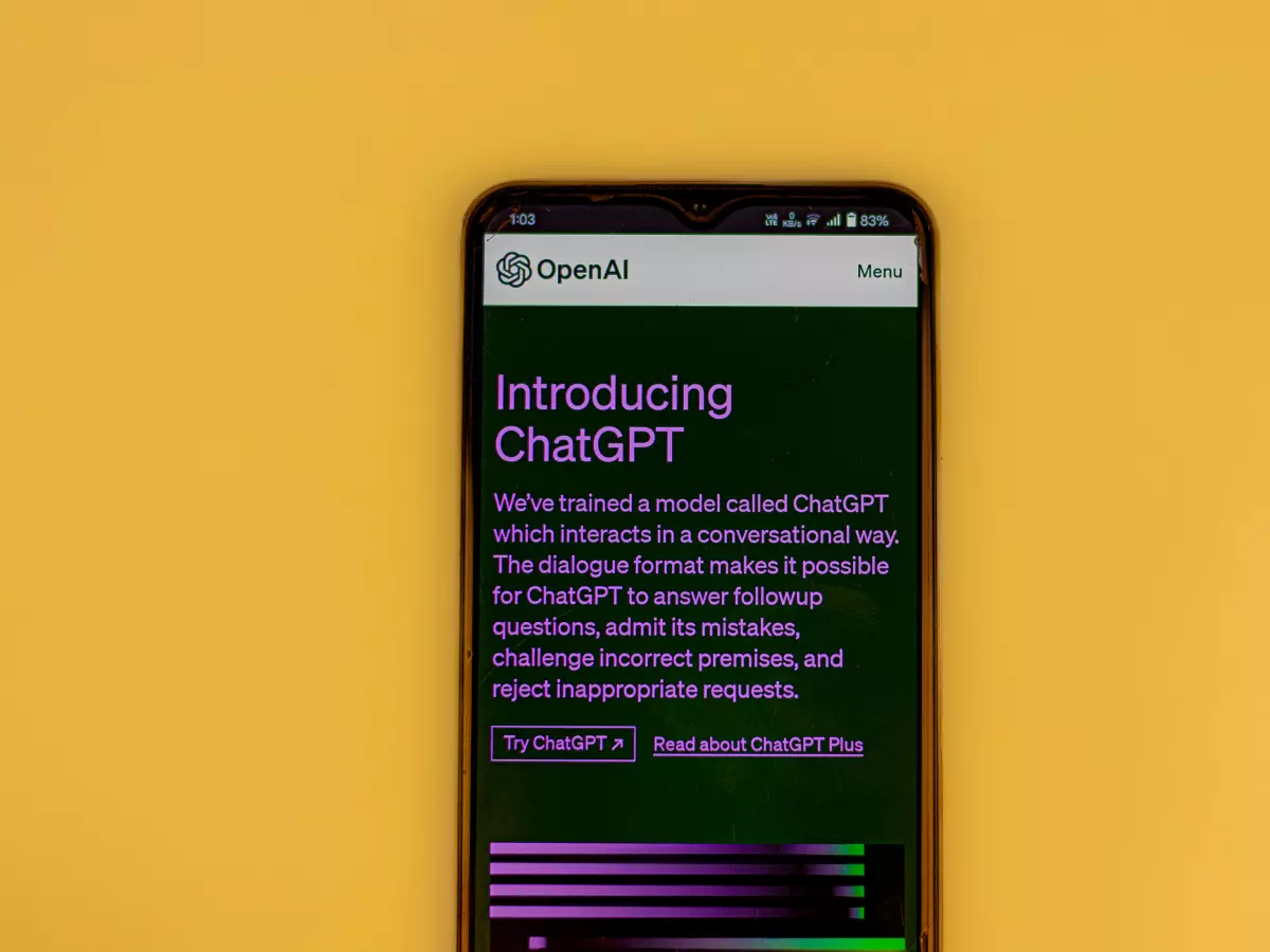 A smartphone with a bright yellow background displaying the website "Introducing ChatGPT" with a description about the service. The text on the phone is in purple and green colors, while the background is black.