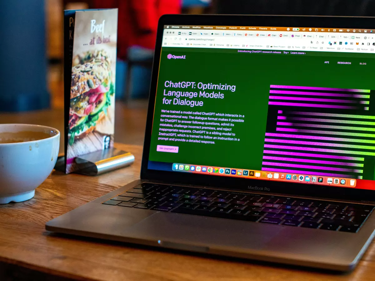 A laptop is open on a wooden table, displaying the ChatGPT website. The text on the screen discusses ChatGPT's optimization of language models for dialogue. A cup of coffee and a menu are visible in the background.