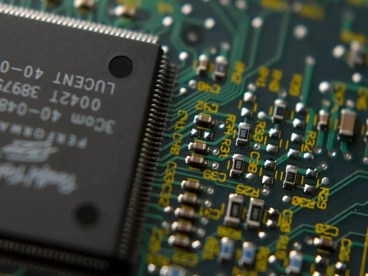 A close-up of a circuit board with a chip in the center. The chip is a small black rectangle with gold pins on the sides. The circuit board is green and has many other components, including resistors, capacitors, and inductors.