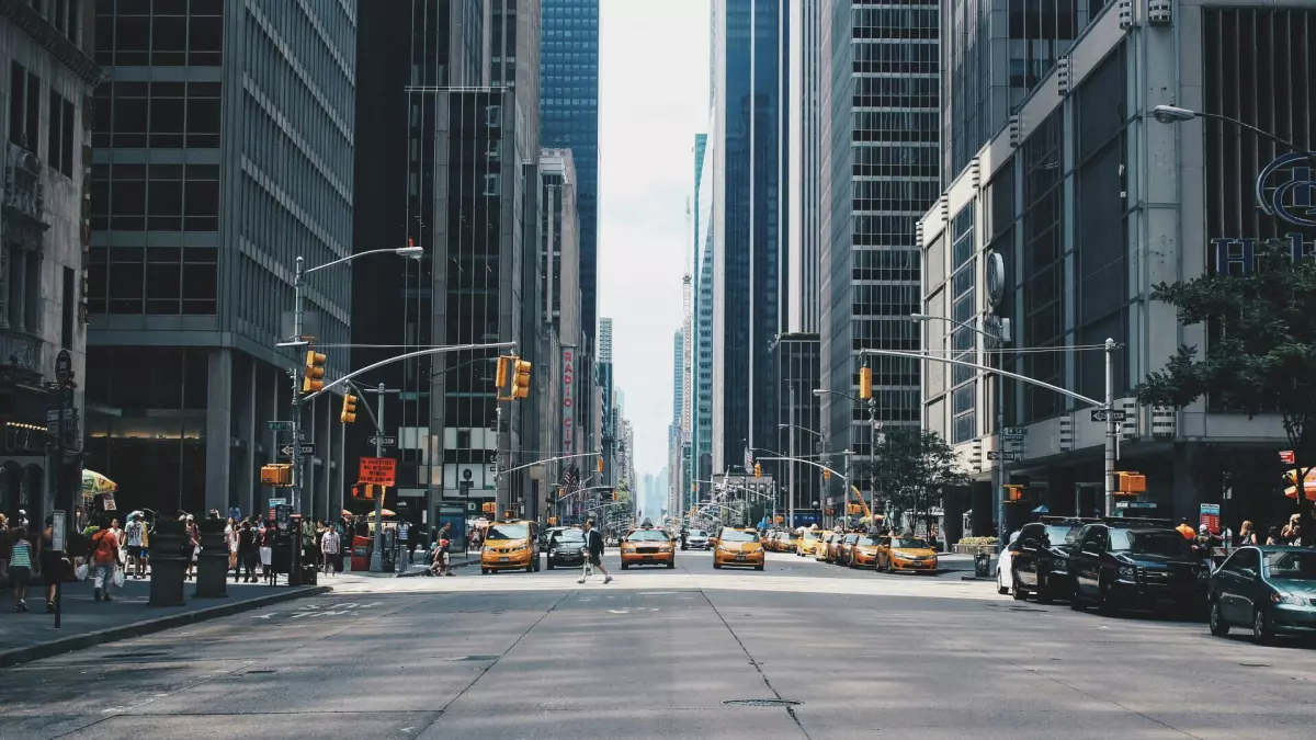 A wide shot of a busy street in a modern city, with tall buildings, parked cars, and pedestrians.