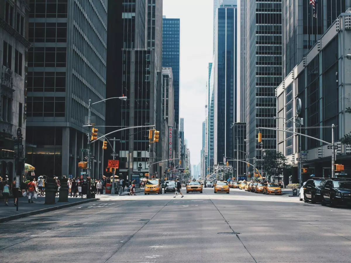 A wide shot of a busy street in a modern city, with tall buildings, parked cars, and pedestrians.