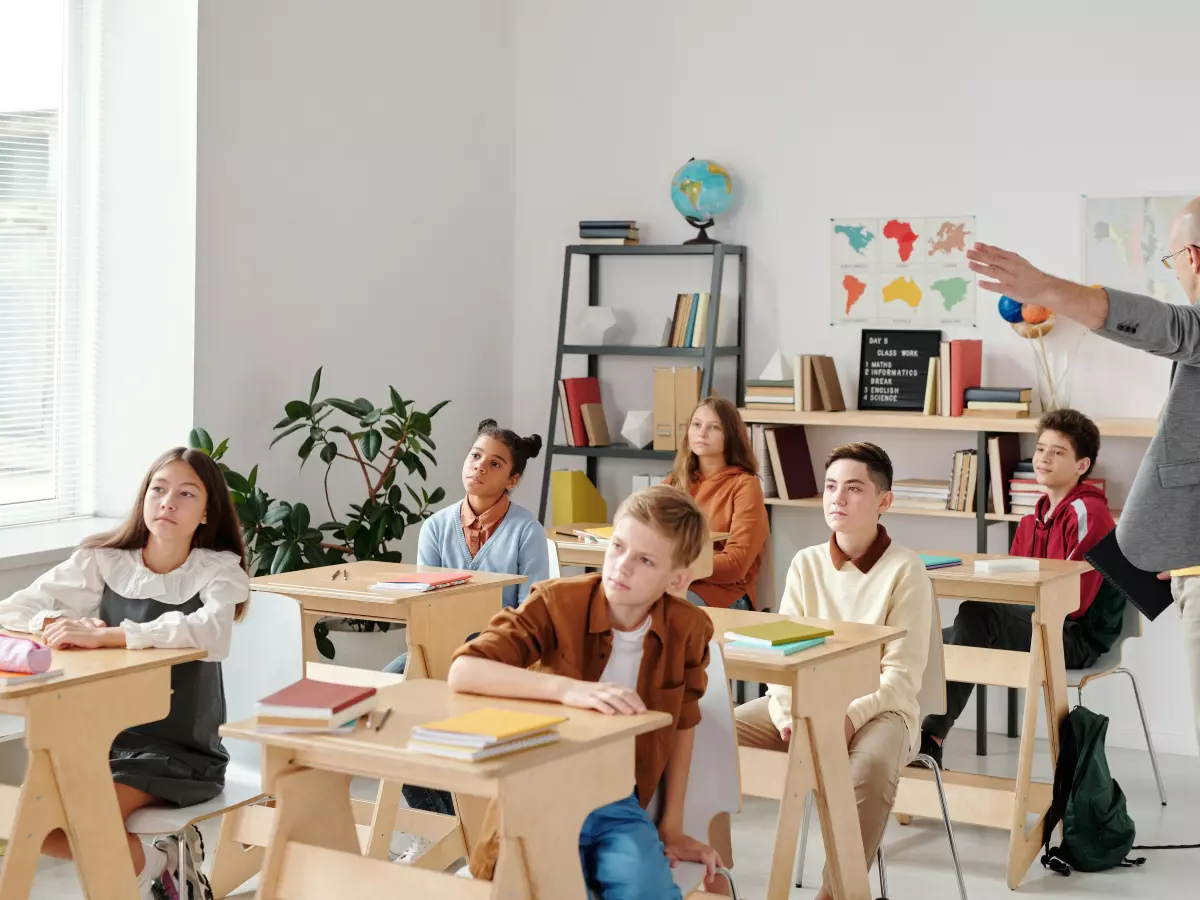 A modern classroom with students listening to their teacher. The teacher is standing and pointing to a board. The students are sitting at their desks, using laptops. There are colourful notes and charts on the wall. The image is bright and airy, showing the promise of AI-powered classrooms.