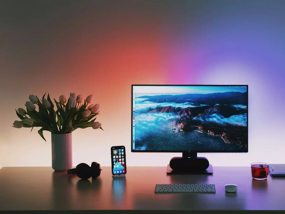 A sleek, modern desk setup featuring a computer monitor with vibrant, colorful lighting behind it. The monitor displays a scenic image, and a smartphone, headphones, and a laptop are visible on the desk.