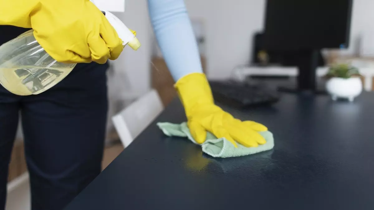 A person wearing yellow gloves is cleaning a desk with a green cloth and a spray bottle. The image is taken from a low angle, showing the person