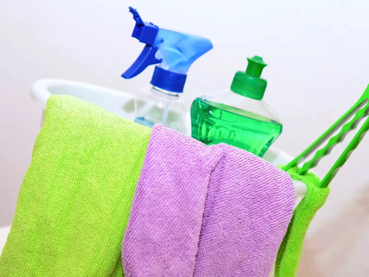 A bucket of cleaning supplies, including a spray bottle, a bottle of dish soap, a green sponge, and two cloths.