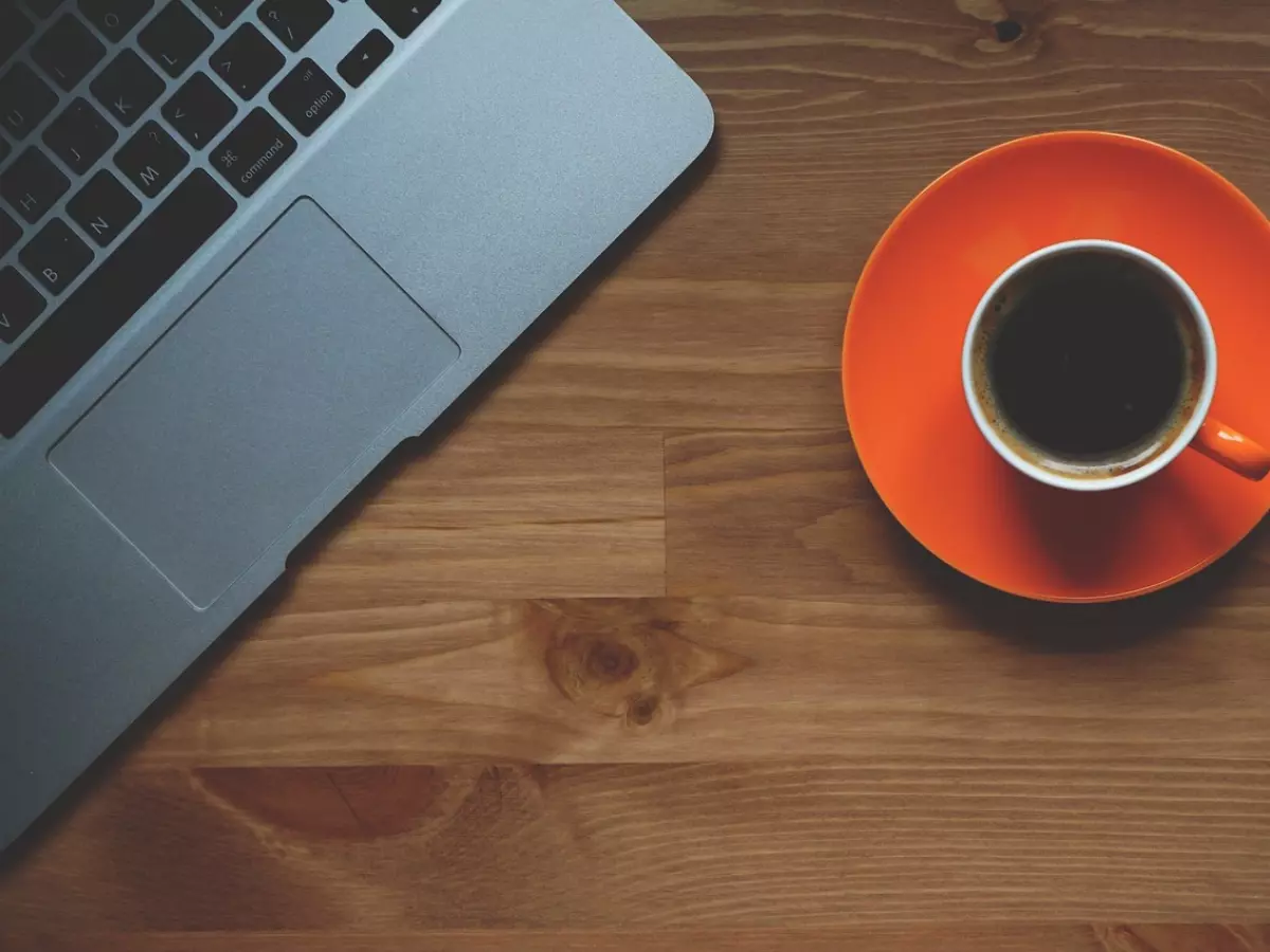 A top-down view of a wooden table with a laptop in the upper left corner and a cup of coffee on a bright orange saucer in the lower right corner. The laptop is open, with its keyboard and trackpad visible, and the coffee cup is full of dark coffee. The wooden table has a natural wood grain and is illuminated by warm, indirect lighting. The image evokes a sense of calm and productivity.