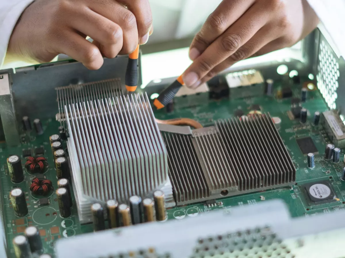 A person is working on a green circuit board with a soldering iron and a pair of tweezers. The image is taken from a close-up perspective. 