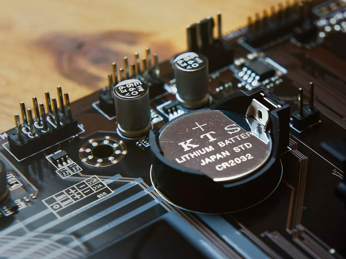 A macro shot of a lithium battery mounted on a circuit board. The battery is small and round, with a black casing and a white label indicating 'KTS Lithium Battery Japan STD CR2032'. The surrounding circuit board is brown and has various components.