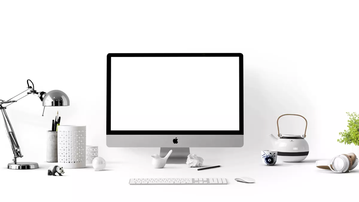 A white desk with a silver computer, a white keyboard and mouse, a white teapot and cup, a lamp, a panda figurine, and headphones.