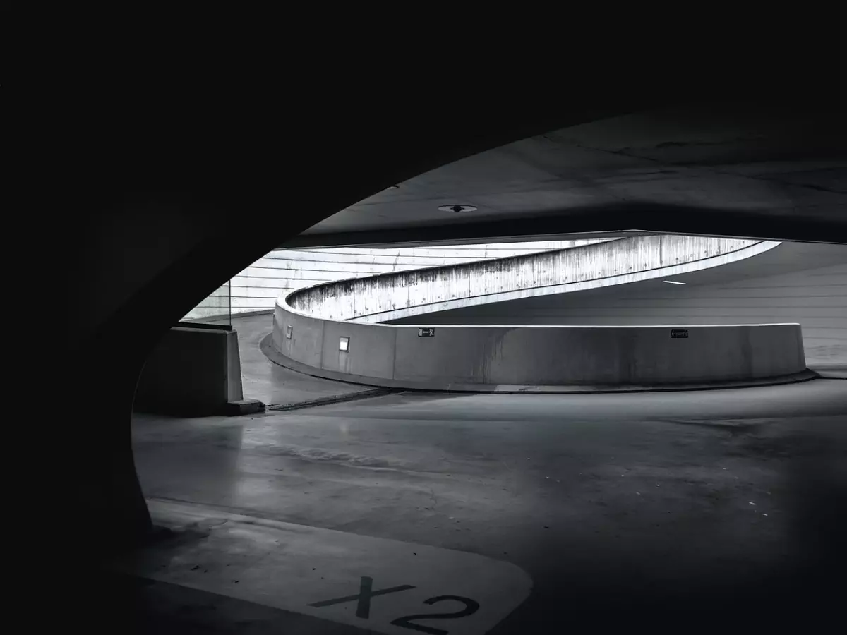 An abstract black and white photo of a modern, curved concrete structure. The image is taken from the inside of the structure, looking out towards a bright, open space.