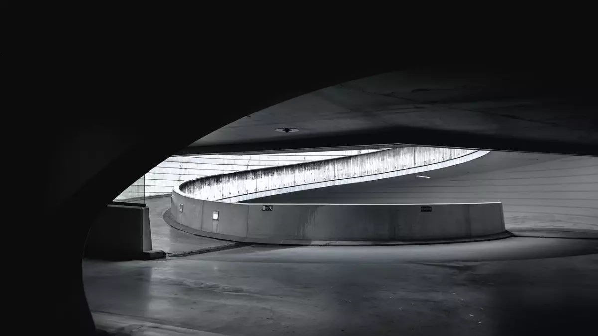 An abstract black and white photo of a modern, curved concrete structure. The image is taken from the inside of the structure, looking out towards a bright, open space.