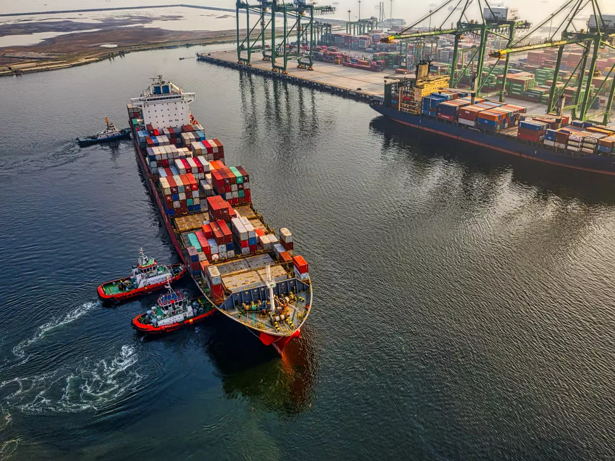 Aerial view of a large container ship being assisted by tugboats in a harbor.