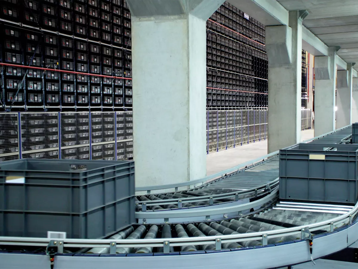 A modern warehouse interior with a conveyor belt system and stacks of crates.  The conveyor belt is moving in the foreground, with crates in different sizes on it. The background shows rows of racks with stacks of crates.