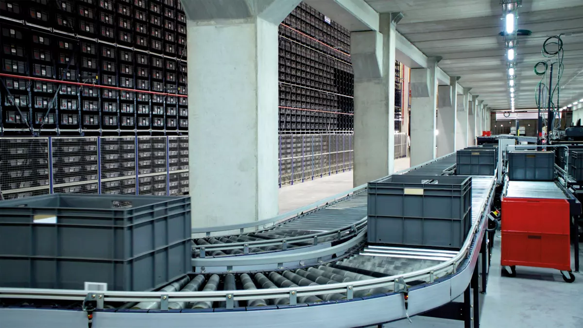 A modern warehouse interior with a conveyor belt system and stacks of crates.  The conveyor belt is moving in the foreground, with crates in different sizes on it. The background shows rows of racks with stacks of crates.