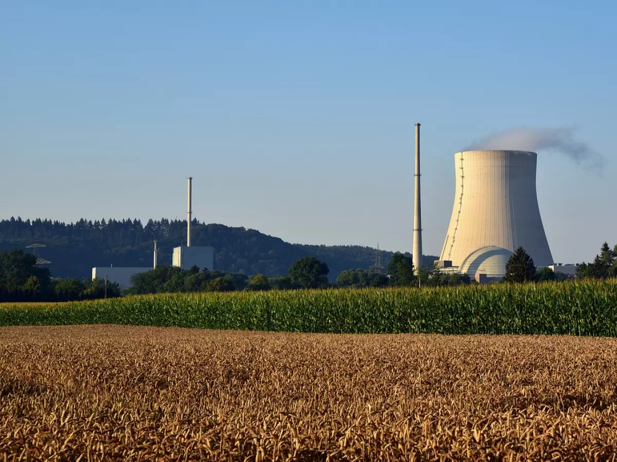 A wide shot of a nuclear power plant with two cooling towers and a large plume of steam rising from the chimney. The plant is set in a rural landscape with fields of corn and wheat in the foreground.