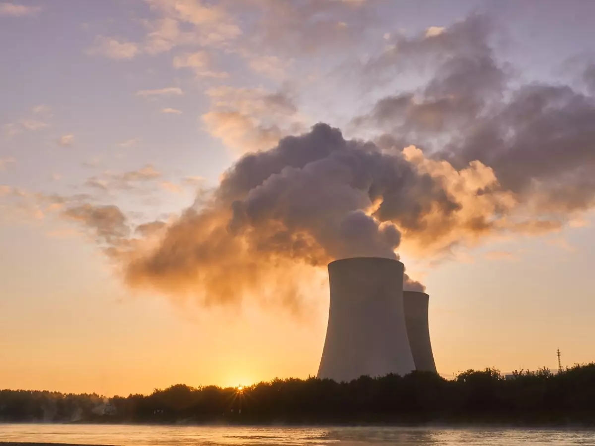 A dramatic sunrise over a nuclear power plant. The image shows two large cooling towers emitting steam into the air, with a river in the foreground.