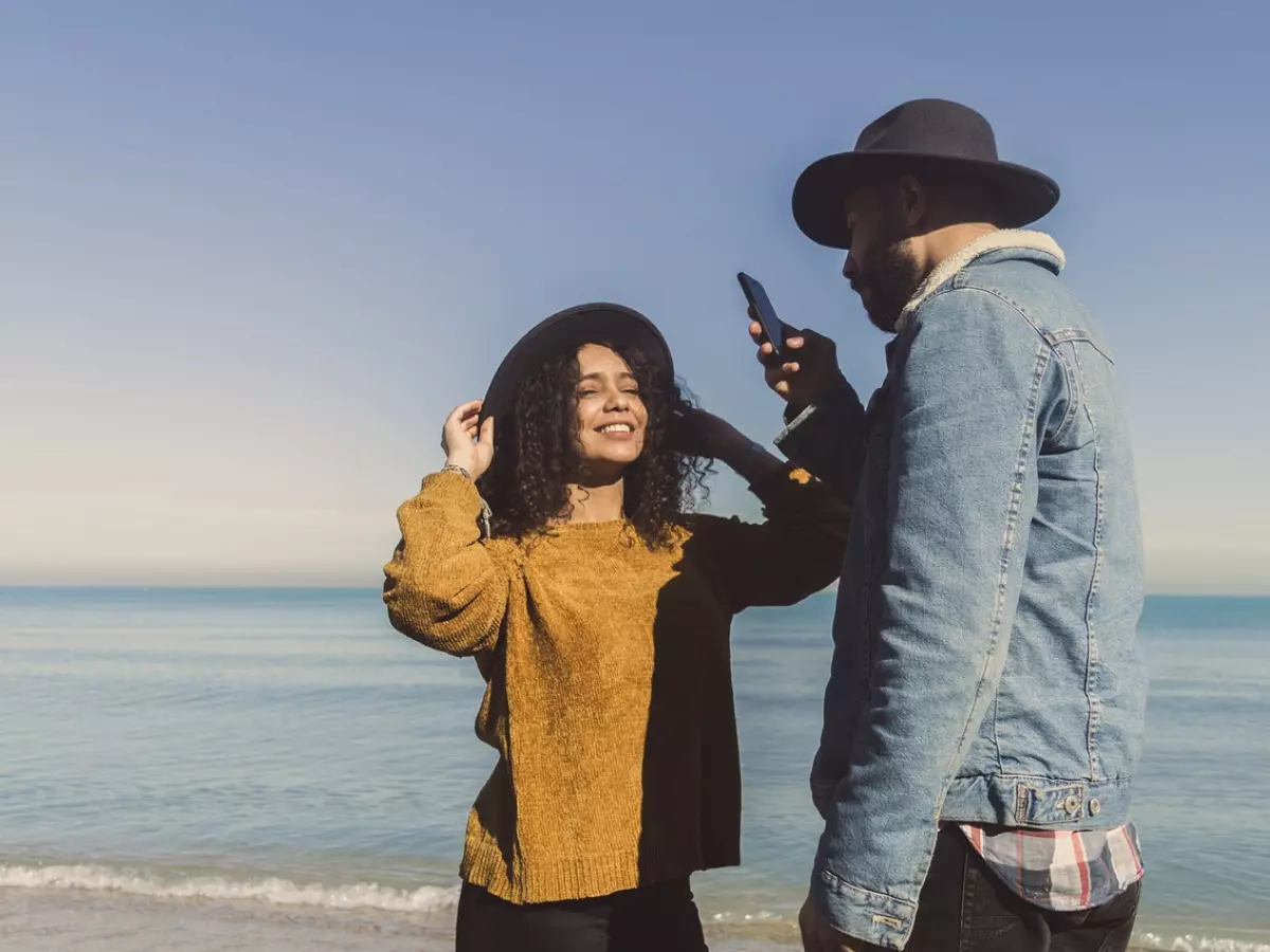 Two people are looking at a phone. The woman is adjusting her hat. They are on a beach.