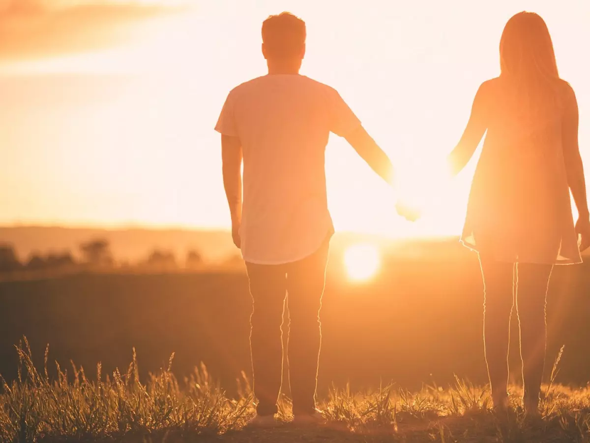 A couple is holding hands, standing in a field with their backs to the camera. The sun is setting in the background, casting a warm glow over the scene.