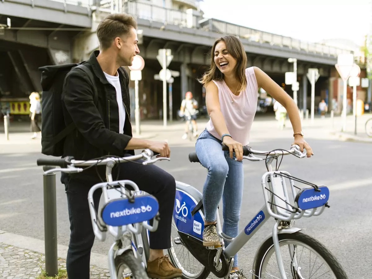 Two people on bikes are smiling and looking at each other, they are both holding smartphones.