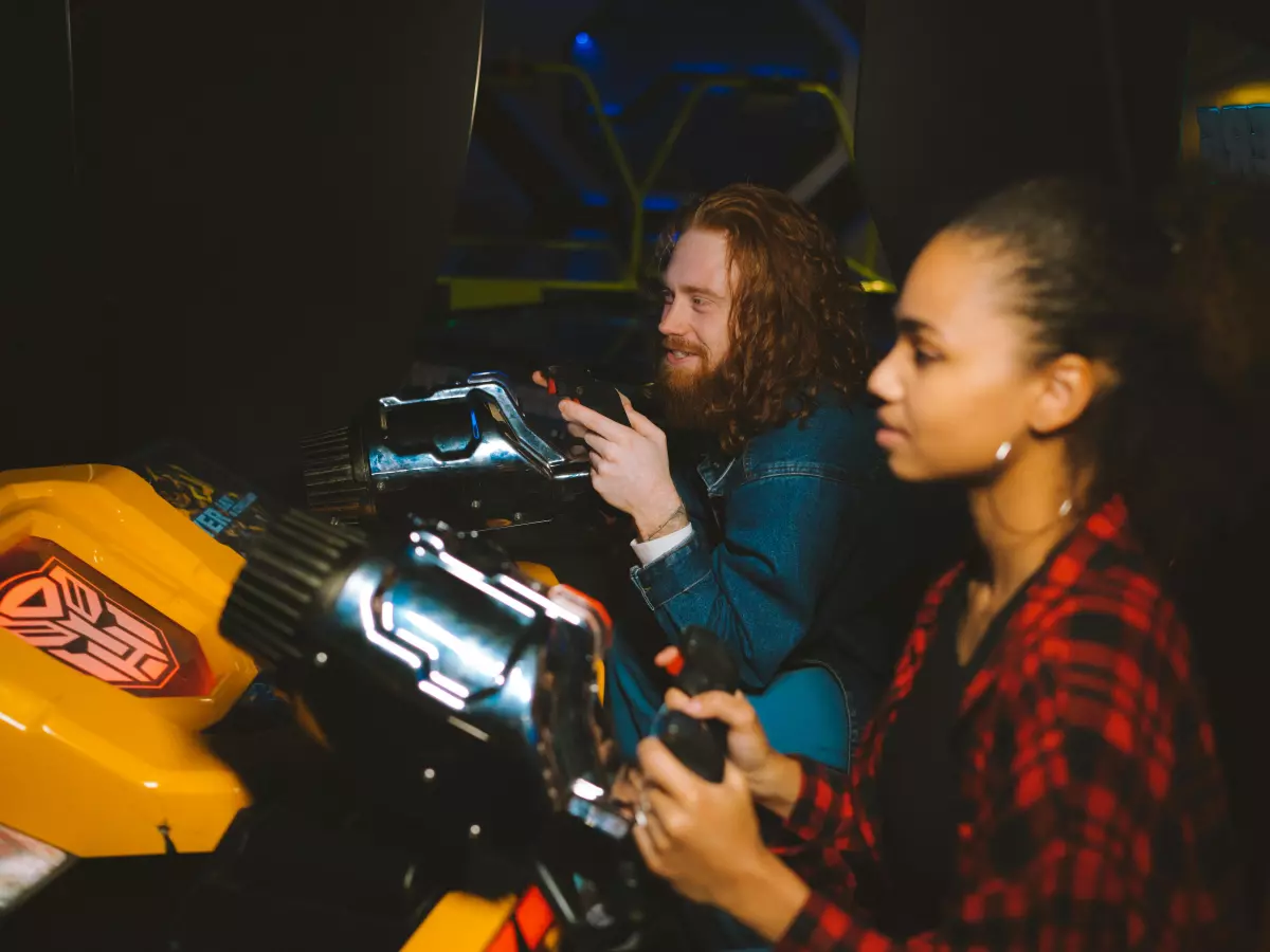 A young woman with a red and black plaid shirt is playing a video game while sitting in a race car simulator, her friend is in the driver's seat. The game is projected on a screen behind them, but you can only see the front part of the car in this photo. The background is dark and mysterious, highlighting the couple and their fun experience.