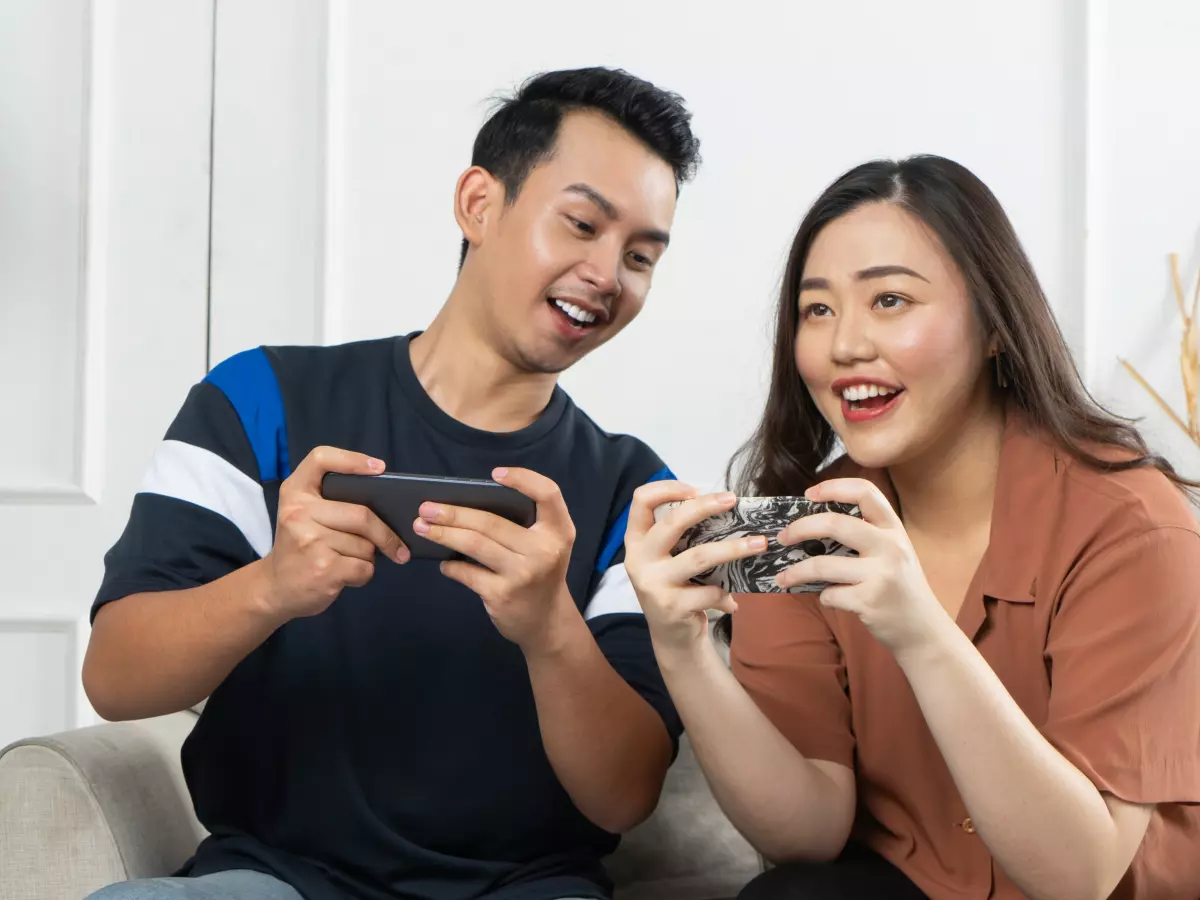 A couple sits on a couch, both playing a game on their phones. 