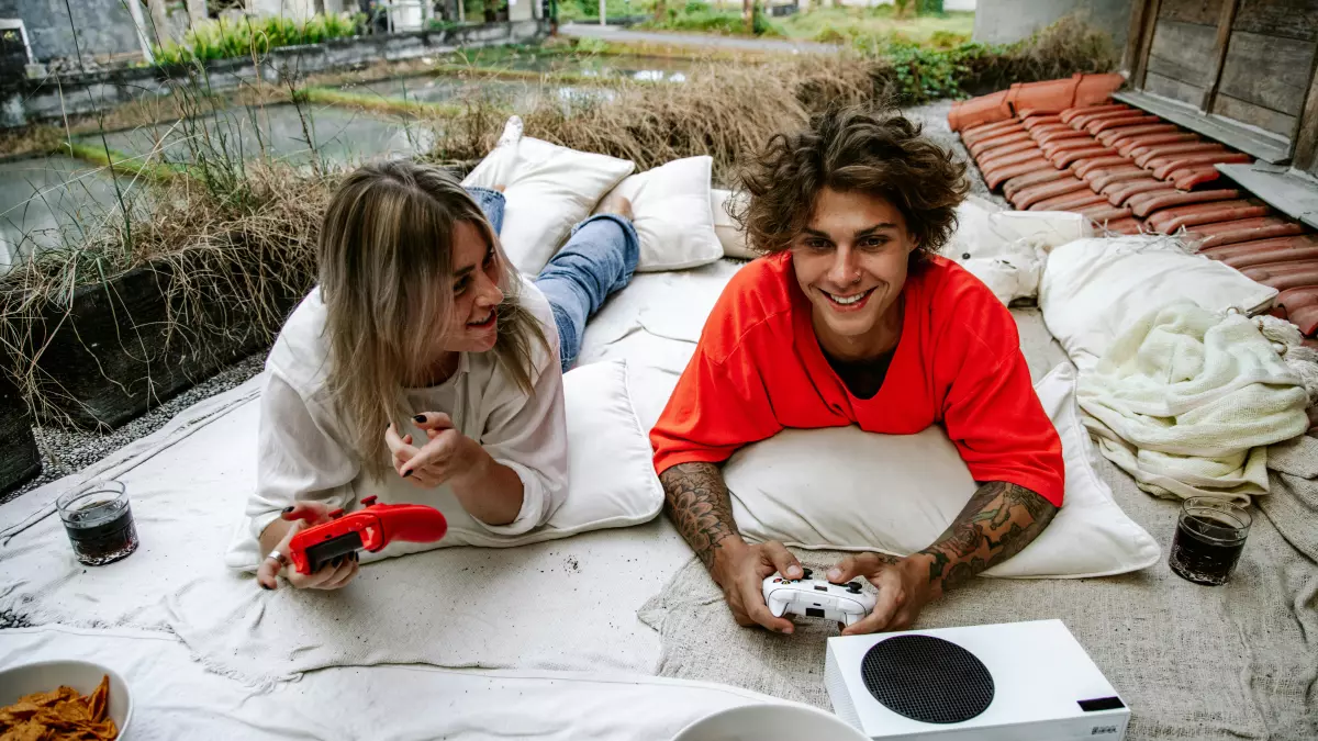 A couple is laying down on a patio, playing video games on a white PS5 console. The man is in a red shirt and the woman is in a white shirt.  They are smiling and looking at each other. There are snacks and drinks on the patio.